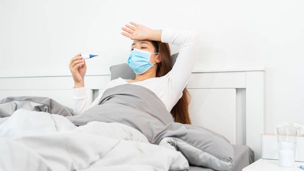 Zieke Aziatische vrouw met beschermend masker liggend in bed met hoge koorts en meten lichaamstemperatuur door thermometer en aanraken van haar voorhoofd bij haar huis - Foto, afbeelding