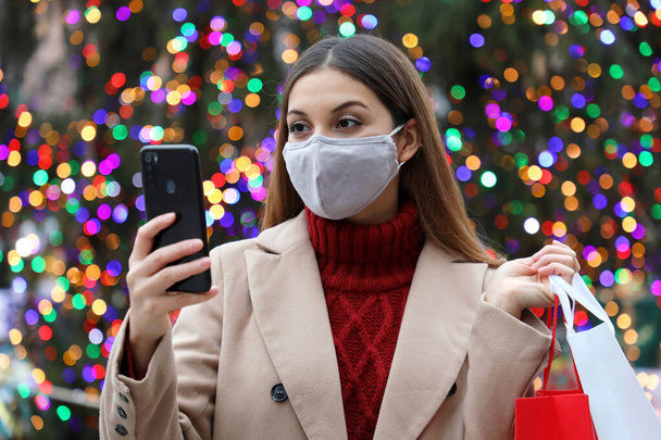 Świąteczna dziewczyna. Szczęśliwa młoda kobieta z maską na twarz niosąca torby na zakupy w ręku kupująca prezenty świąteczne ze smartfonem na świeżym powietrzu. - Zdjęcie, obraz
