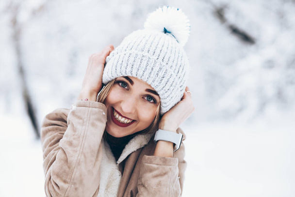 νεαρή γυναίκα σε πλεκτό καπέλο χαμογελώντας στο χιονισμένο πάρκο - Φωτογραφία, εικόνα