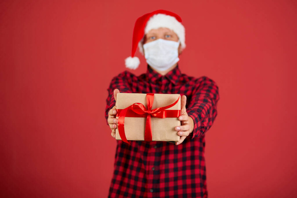 Man in protective face mask and Santa hat with Christmas DIY gift during covid καραντίνα, Coronavirus νέο έτος concept, courier παράδοση στο σπίτι. - Φωτογραφία, εικόνα