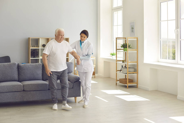 Friendly nurse helps an elderly patient walk around the room in a nursing home holding his hand. - Foto, afbeelding