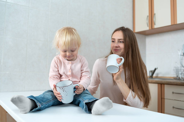 Szczęśliwa młoda matka i córka siedzą przy drewnianym stole w kuchni i trzymają białe kubki. - Zdjęcie, obraz