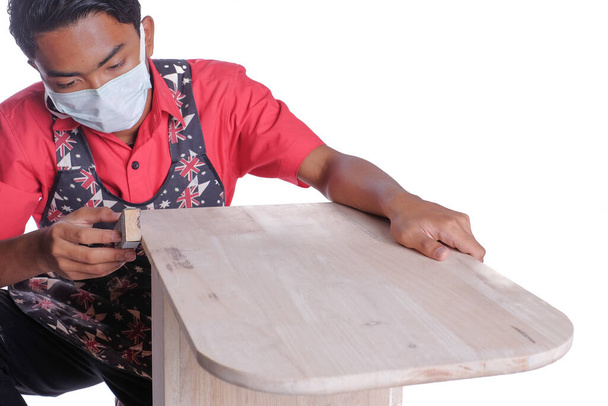 Jeune menuisier en vêtements de travail en utilisant du papier de verre pour le projet de serveur de canapé de polissage isolé sur fond blanc - Photo, image