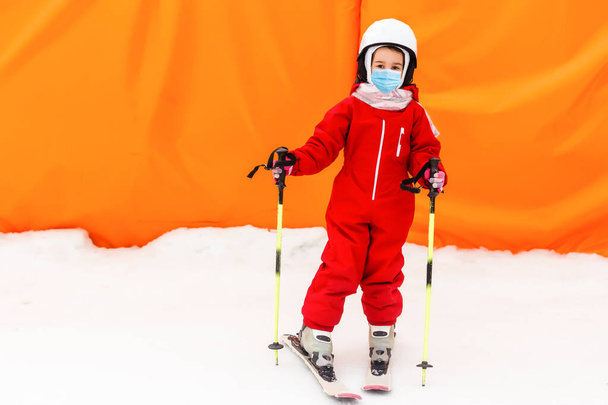 Porträt eines kleinen Skifahrers mit medizinischer Maske während des COVID-19 Coronavirus auf einem schneebedeckten Berg in einem Skigebiet - Foto, Bild