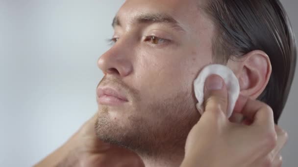 Primo piano colpo di irriconoscibile make-up artist maschile utilizzando batuffoli di cotone e strisciandoli sul viso di calmo giovane uomo per pulire la pelle - Filmati, video