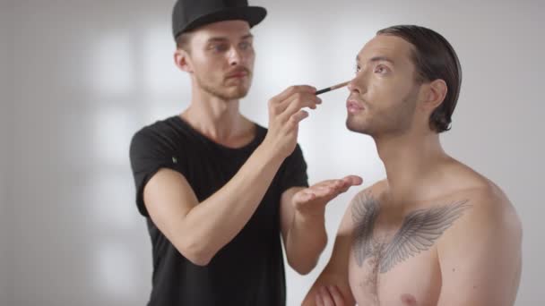 Keskikokoinen laukaus ammatillinen mies meikkitaiteilija käyttäen harjaa ja soveltamalla kosmeettinen valmiste silmän alla nuori mies studiossa - Materiaali, video