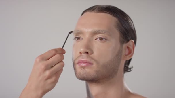 Lähikuva kädestä tunnistamaton meikkitaiteilija käyttäen spoolie kammata läpi kulmakarvat komea nuori mies - Materiaali, video
