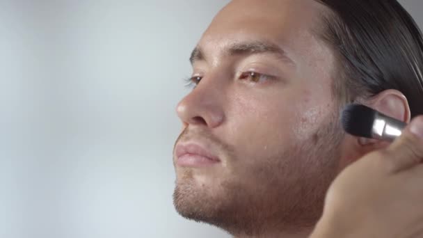Detailní záběr nerozpoznatelné make-up umělce pomocí štětce a nanášení základní nebo hydratační krém na tvář mužského modelu - Záběry, video