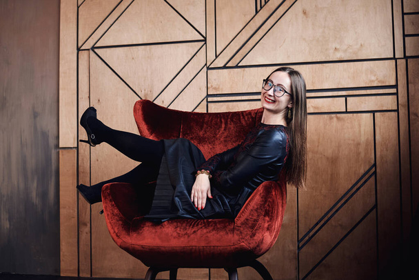 Young brunette woman, wearing fancy black leather dress and eyeglasses, sitting on red velvet Santa armchair in front of wooden wall, Merry Christmas and Happy New year celebration. Female portrait - Фото, изображение