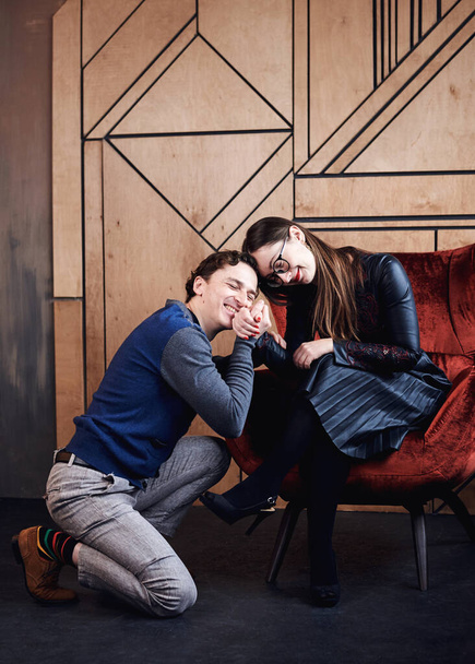 Young woman, sitting on red velvet Santa armchair and man, kneeling down near her in front of wooden wall, smiling, laughing. Romantic relationship. Young couple in fancy clothes, posing for picture. - Foto, afbeelding