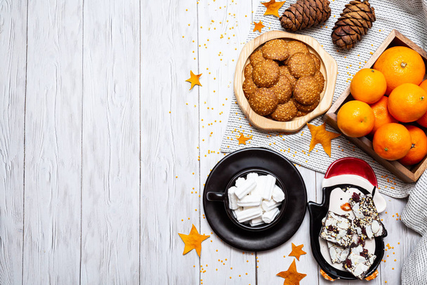 Cozy winter still life with tangerines, cookies, chocolate, pine cones and a cup of marshmallows on a light wooden table. Top view, place for text. - Photo, Image