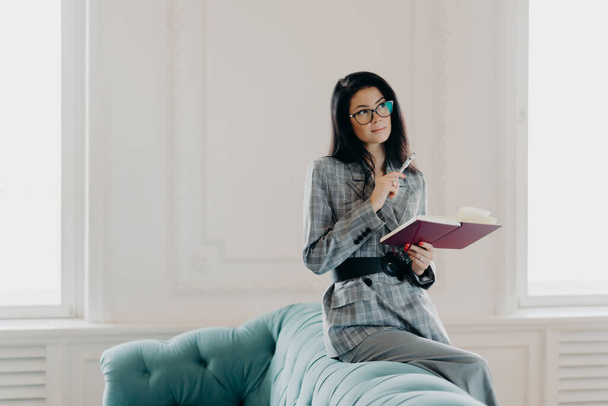 Serious businesswoman dressed in formal wear, concentrated into distance, ponders on creative ideas, writes down notes in notepad, wears spectacles, poses on back of couch in spacious light room - Fotografie, Obrázek