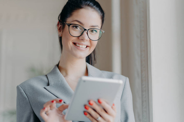 Vnitřní záběr šťastné brunetky podnikatel čte elektronickou knihu na moderním digitálním tabletu, čte finanční zprávy na internetu během pracovní přestávky, nosí optické brýle, šedý kostým, pózuje uvnitř - Fotografie, Obrázek