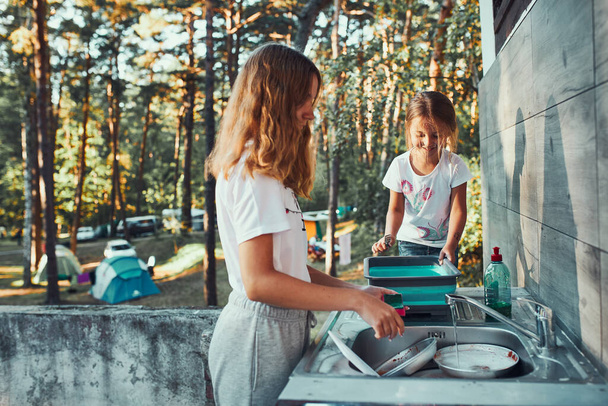 Teenager girl washing up the dishes pots and plates with help her younger sister in the outdoor kitchen during vacations on camping. Camp life. Sisters spending time and working together - Foto, afbeelding