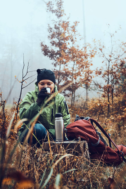 Woman with backpack having break during autumn trip drinking a hot drink from thermos flask on autumn cold day. Active middle age woman wandering around a forest actively spending time - Foto, Imagem