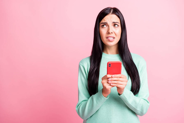 Photo portrait of ashamed girl biting lower lip looking at blank space holding phone with two hands isolated on pastel pink colored background - Fotó, kép
