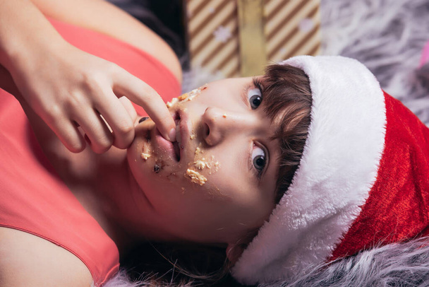 Portrait of a teenage girl in a santa hat with a dirty face in sweet cream from Christmas cakes. High quality photo - Foto, immagini