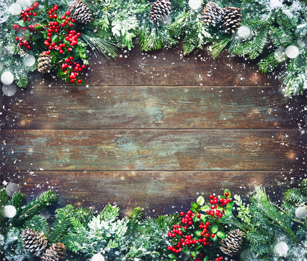 Christmas and New Year background with fir branches, holly and snowfall on dark wooden board - Photo, image