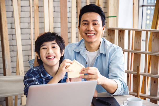 Küçük ahşap ev modelini tutan mutlu Asyalı baba ve oğul marangozluk atölyesinde dizüstü bilgisayar kullanıyorlar. Aile mesleği hobisi.. - Fotoğraf, Görsel
