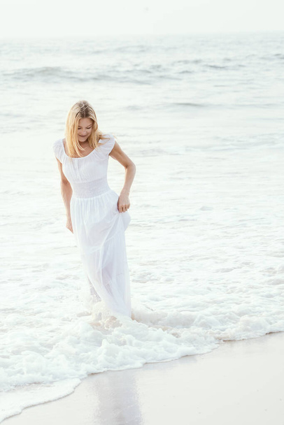 アジアの砂の熱帯のビーチで長い白いドレスを着た魅力的な幸せな成熟した観光客のブロンドの女性は、水の近くに喜び笑みを浮かべて。ロマンチックな旅行休暇、コピースペース付きのアクティブなライフスタイル. - 写真・画像