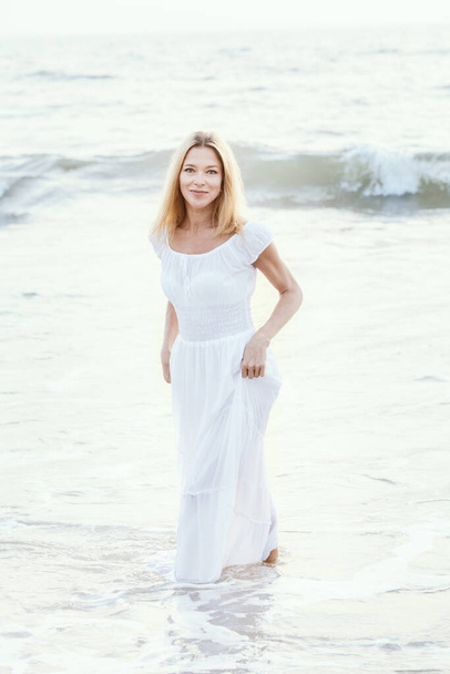 アジアの砂の熱帯のビーチで長い白いドレスを着た魅力的な幸せな成熟した観光客のブロンドの女性は、水の近くに喜び笑みを浮かべて。ロマンチックな旅行休暇、アクティブなライフスタイル、コピースペースを持つ独立した自信のある人. - 写真・画像