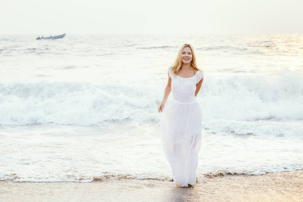 Attractive happy mature tourist blonde woman in long white dress on asian sand tropical beach smiling joyful near water. Romantic travel vacation, active lifestyle, independent and confident person with copyspace. - 写真・画像