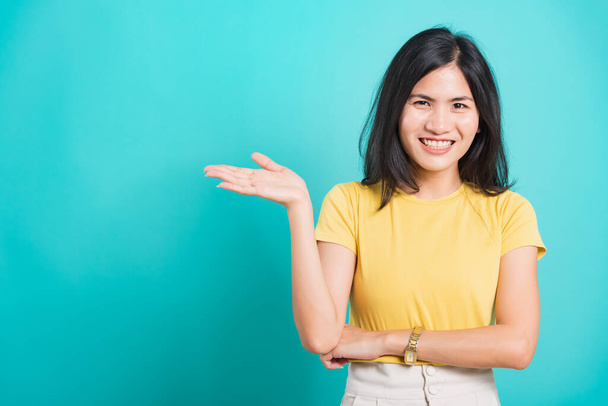 Portrait Asian beautiful young woman standing wear yellow t-shirt, She showing hand to presenting product and looking at the camera, shoot photo in a studio on a blue background, There was copy space - 写真・画像