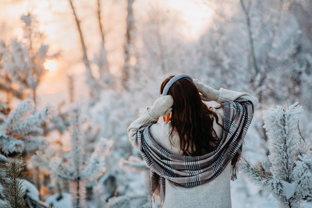 Девушка в бежевых клетчатых и меховых наушниках веселится в сказочном снежном лесу. Снежные деревья. Модная стильная теплая зимняя одежда. Искусственный мех. Время заката - Фото, изображение