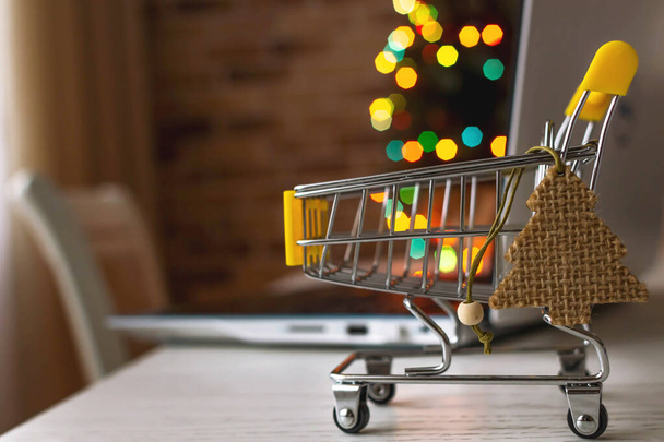 Christmas shopping online. Laptop and shopping cart at home on the background of a Christmas tree with blurred bokeh lights. - Φωτογραφία, εικόνα