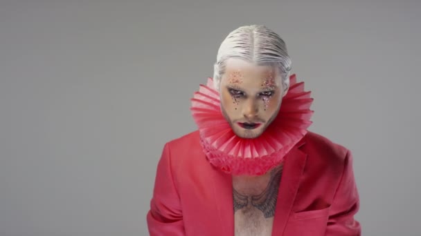 Portret mężczyzny z dramatycznym makijażem z brokatem i wyszczuplonymi białymi włosami z czerwonym rufą na szyi i kurtką na gołej klatce piersiowej, pozujący do kamery - Materiał filmowy, wideo