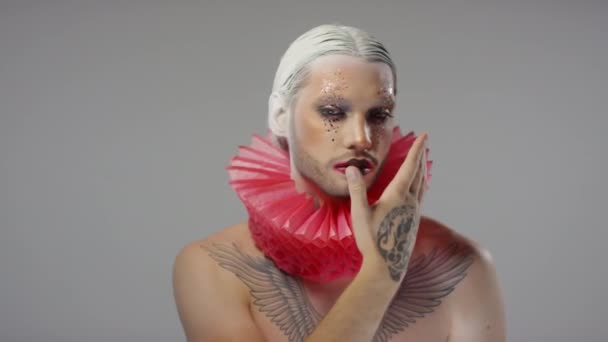 Πορτρέτο του κομψού νέου άνδρα με θεατρικό μακιγιάζ, κόκκινο ρούφηγμα γύρω από το λαιμό του και φέτες πίσω άσπρα μαλλιά ποζάρουν σε γκρι φόντο - Πλάνα, βίντεο