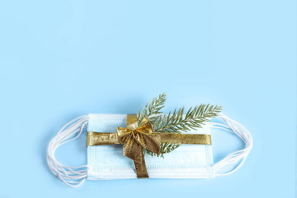 金色のリボンと青の背景に隔離された枝モミの木と医療保護マスク。クリスマスと新年のための贈り物2021 。コロナウイルスパンデミックの概念。スペースのコピー. - 写真・画像
