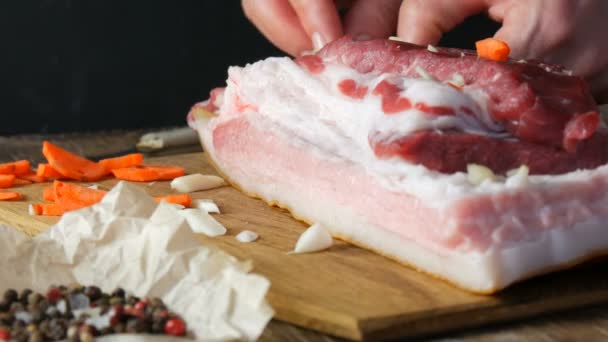 Vrouwelijke handen steken met een mes en gevulde kruiden met plakjes knoflook en wortelen een stuk vers rauw vlees varkensvlees steak of spek in een rustieke stijl op houten keukenbord. - Video