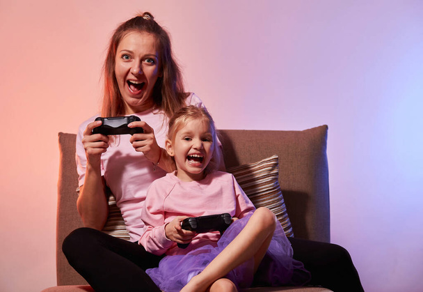 Emocionada madre joven y linda hija pequeña se sientan en el sofá sosteniendo el mando del mando mientras juegan videojuegos en casa juntos, se divierten en la sala de estar con luz de neón azul rojo - Foto, imagen