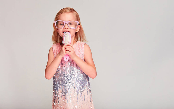 Funny kid girl sing song holding hair brush like a microphone in pink dress and glasses over grey background with copy space. - Foto, afbeelding