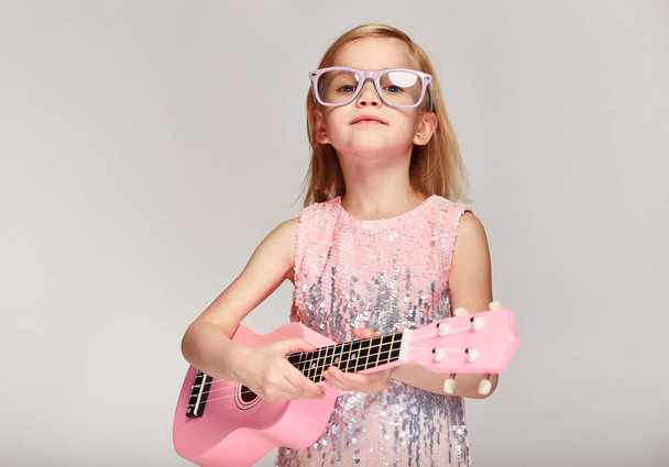 Rock and roll music koncepce.Roztomilý kavkazský holčička 4-5 let v jasně růžové šaty a brýle hrají ukulele na šedém pozadí studia s kopírovacím prostorem - Fotografie, Obrázek