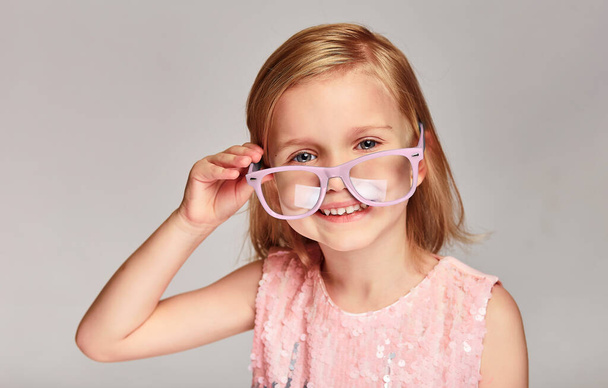 Portrait 4-5 year old kid girl posing in pink glasses and dress over grey background with copy space - Photo, Image