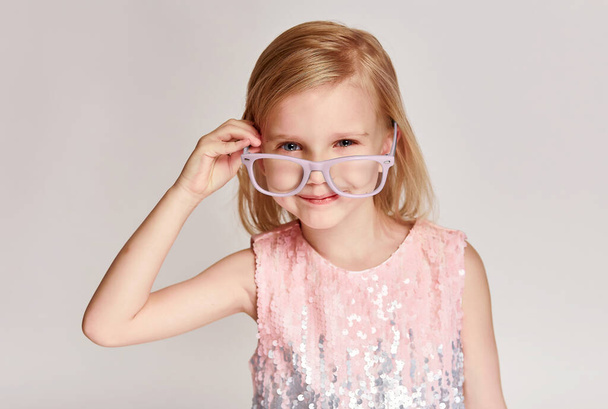 Ritratto bambina di 4-5 anni in posa in occhiali rosa e vestito su sfondo grigio con spazio copia - Foto, immagini