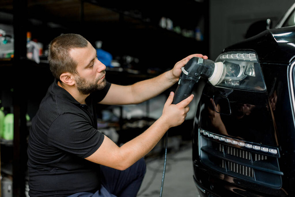 Close up of professional Caucasian male auto service worker, waxing and polishing headlight of black car with orbital polisher. Car detailing series, polishing concept. - Fotó, kép