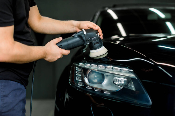 Car polishing process. Cropped image of hands of male ervice employee in working clothes, polishing black car hood with orbital polishing machine, wax ceramic coating - 写真・画像