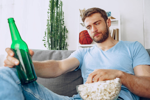 Young bearded man sitting on couch with beer and popcorn while watching movie - Fotografie, Obrázek