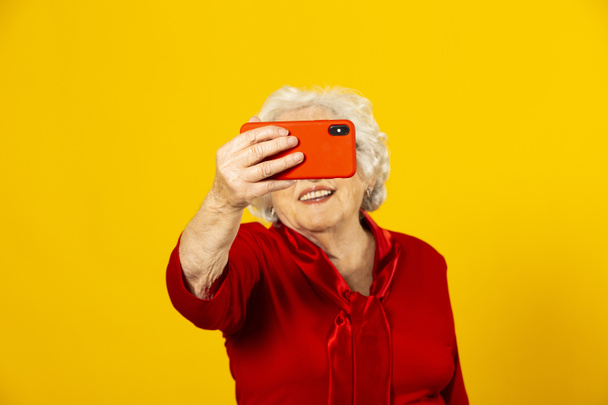 黄色の背景に赤いシャツを着て、赤い携帯電話でビデオ通話をしているシニア女性のスタジオの肖像画 - 写真・画像