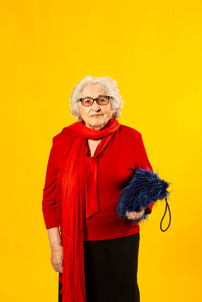 Студийный портрет пожилой женщины в красной рубашке и синей волосатой сумке на жёлтом фоне - Фото, изображение