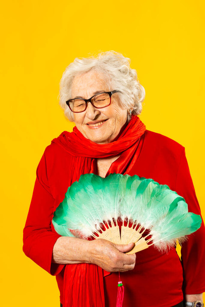 Studio portrait of a senior woman wearing a red shirt and a green feather hand fan, against a yellow background - Foto, Imagen