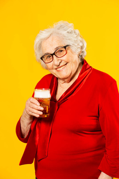 Studio portrait of a senior woman wearing red shirt with a glass of a half pint beer, against a yellow background - Photo, Image