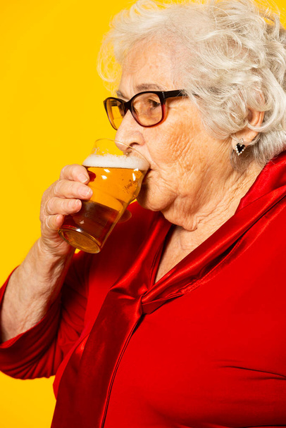 Kırmızı tişört giyen, sarı arka planda yarım litre bira içen son sınıf bir kadının stüdyo portresi. - Fotoğraf, Görsel