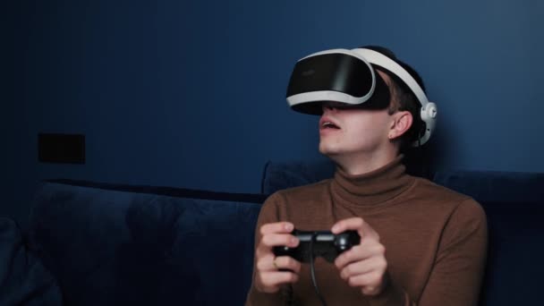Ελκυστικός καυκάσιος άντρας κρατά χειριστήριο και φοράει γυαλιά εικονικής πραγματικότητας κράνος κεφαλής σε καναπέ στο σπίτι. Ευτυχία gamer παιχνίδι παίζοντας χρησιμοποιώντας γυαλιά VR στο σπίτι. - Πλάνα, βίντεο