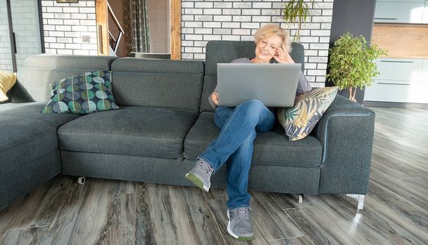 高齢者のかなりの女性がノートパソコンでリビングルームのソファに座っています。友人や親戚とのオンライン通信をリモートで。女性はビデオ会議でお祝いを受け入れる. - 写真・画像