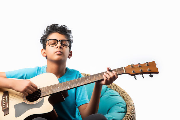 Indian asian boy playing acaustic guitar while sitting against white background or brick wala on chair - Φωτογραφία, εικόνα