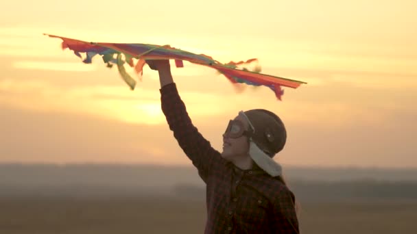 happy child in a pilots helmet runs with a multi-colored kite in his hands at sunset. girl wants to become pilot. child dreams of freedom, flight and travel. teenager playing on a plane outdoors. - Footage, Video